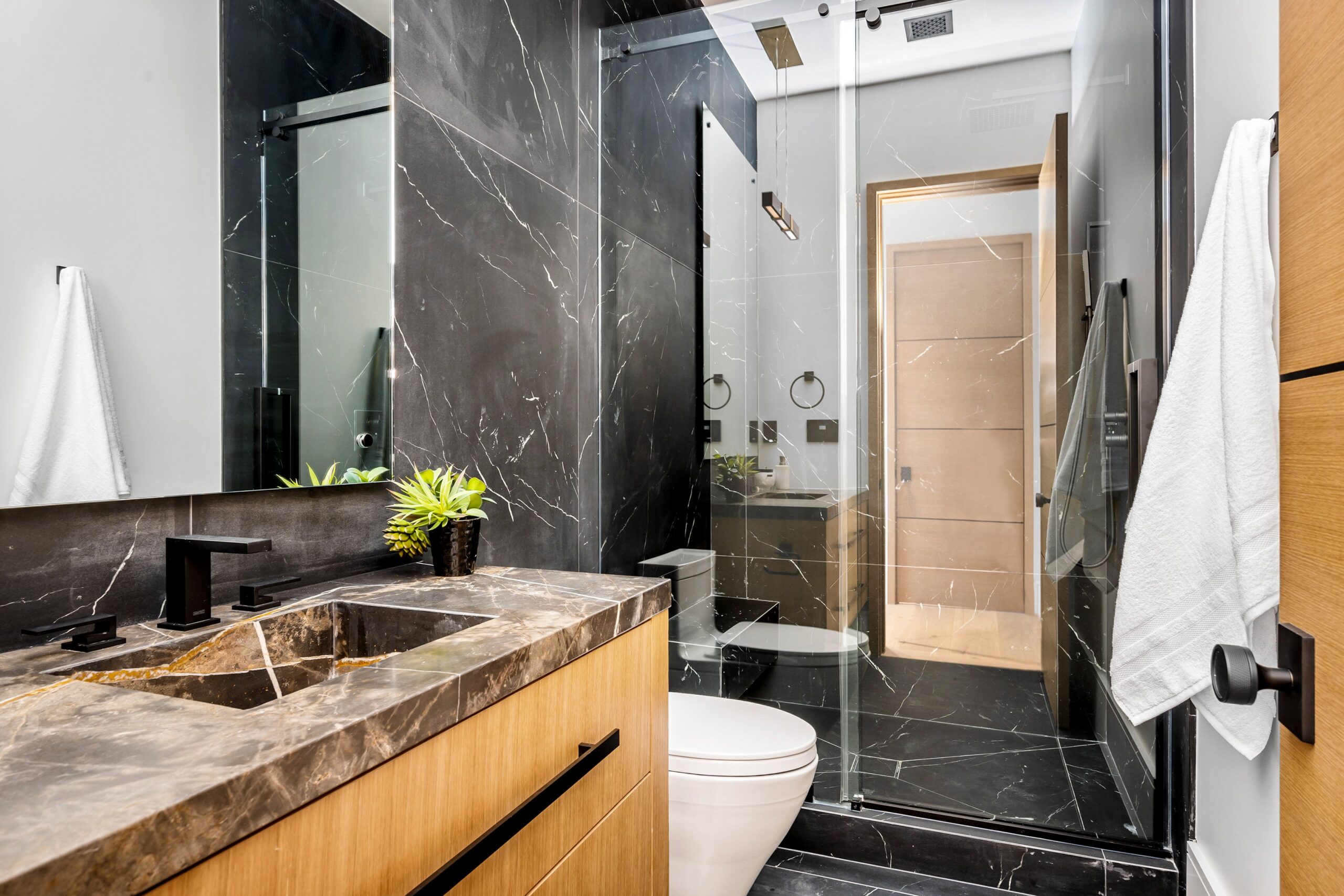 A modern bathroom with black marble tiles, wooden cabinetry, and glass shower doors.